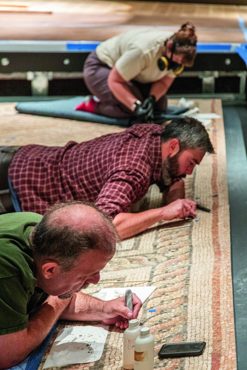 Three people working on the ground, inspecting small pieces of a large mosaic they are installing.